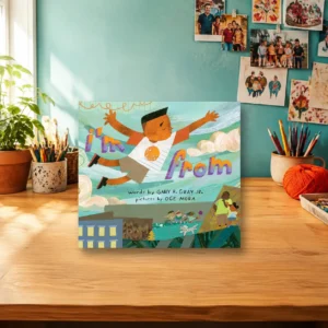 Long Childhood - Children’s book titled “I’m From” is displayed upright on a wooden desk, surrounded by art supplies, plants, and family photos on a teal wall in a well-lit room.